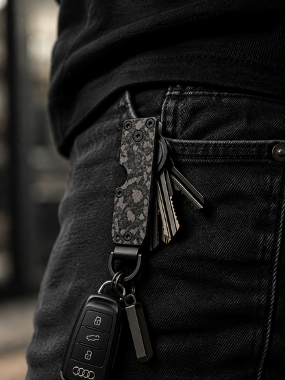 A close-up lifestyle shot of a person's waist wearing black jeans and a black shirt, showing a multi-tool keychain clipped to the back pocket. The keychain features a dark gray and black camouflage patterned pocket knife, a black car key fob, and other keys.